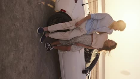 Vertical-video:-A-bearded-man-in-a-blue-shirt-stands-with-his-blonde-girlfriend-in-a-white-top-leaning-on-a-white-convertible-car-against-the-yellow-sky-and-ground