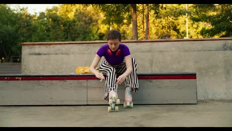 Shooting-from-the-front:-A-young-short-haired-girl-in-a-purple-top-and-striped-pants-lace-up-her-skates-on-a-special-bench-in-a-skate-park-for-skaters