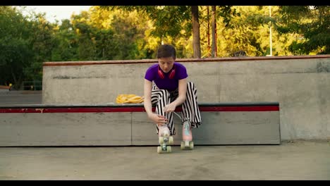 A-young-girl-with-a-short-haircut-in-a-purple-top-sits-on-a-stone-bench-and-laces-up-her-four-wheeled-skates-in-the-park