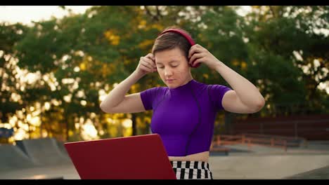 A-young-short-haired-girl-in-a-purple-top-puts-on-red-headphones-and-starts-her-work-with-a-red-laptop-in-the-park-in-summer
