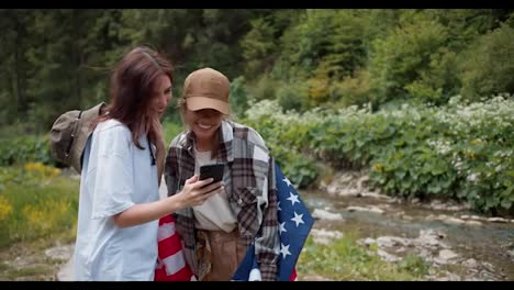 A-brunette-girl-took-a-picture-of-her-friend-with-an-American-flag-And-now-they-are-looking-at-the-photo.-Photo-against-the-background-of-a-green-forest-and-mountain-rivers