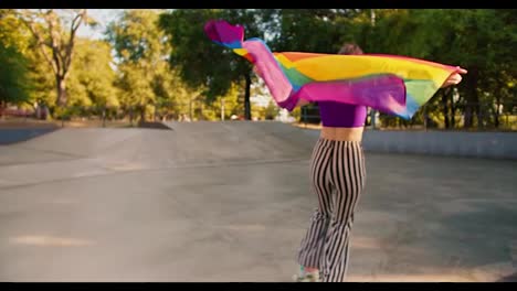 Rückansicht-Eines-Mädchens-In-Einem-Lila-Oberteil-Und-Einer-Gestreiften-Hose,-Das-In-Einem-Skatepark-Rollschuh-Fährt-Und-Eine-Wehende-LGBT-Flagge-Hält