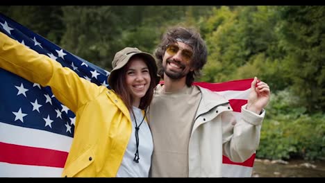 The-brunette-guy-and-girl-pose-and-admire,-they-raised-the-US-flag-during-their-trip-through-the-mountain-forests-against-the-background-of-green-trees