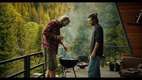 Two-guys-fry-sausages-on-the-grill-on-the-grill.-A-man-in-a-red-shirt-turns-over-sausages-using-special-tongs-against-the-backdrop-of-mountains-and-forest