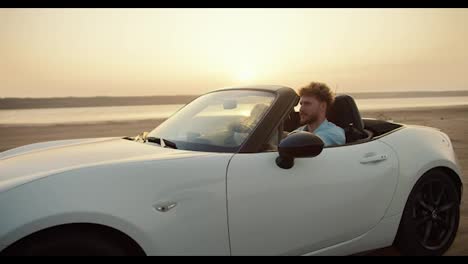 A-bearded-man-with-curly-hair-in-a-blue-shirt-is-driving-with-his-girlfriend-in-a-white-convertible-car-on-the-beach-against-the-yellow-sky