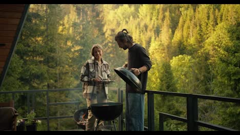 A-young-couple,-a-guy-and-a-girl,-are-relaxing-in-a-country-house-and-frying-meat-on-a-special-grill-against-the-backdrop-of-a-green-coniferous-forest