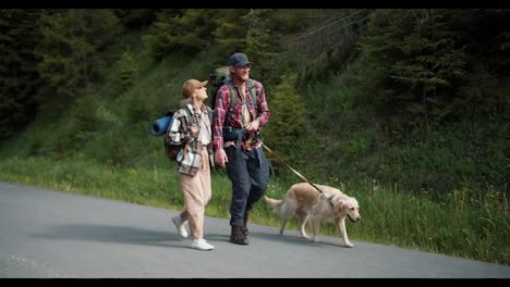 A-blond-guy-in-a-red-shirt-and-a-blond-girl-in-special-clothes-for-hiking-Walk-with-their-dog-Light-colored-along-the-road-along-the-forest-a-They-travel-through-new-territories
