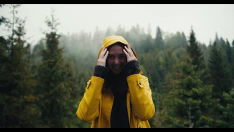 A-happy-girl-in-a-yellow-jacket-stands-against-the-backdrop-of-a-coniferous-mountain-forest,-she-puts-on-a-hood-and-looks-into-the-camera.-It's-raining-a-little