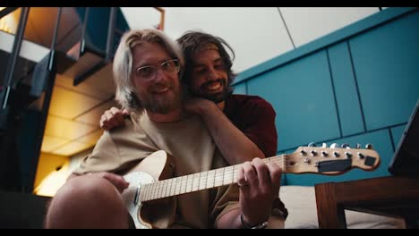Two-guys-friend-spending-time-together-guy-brunette-hugging-guy-blonde-with-a-beard-in-glasses-who-plays-the-electric-guitar-in-a-cozy-room