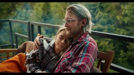 A-girl-hugs-a-blond-guy-with-glasses.-A-couple-is-resting-on-a-devan-wrapped-in-a-blanket-on-the-balcony-of-a-country-house-overlooking-the-mountains-and-forest