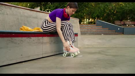 A-young-short-haired-girl-in-a-purple-top-and-striped-pants-laces-up-roller-skates-on-a-skateboarding-site-in-the-park-in-summer