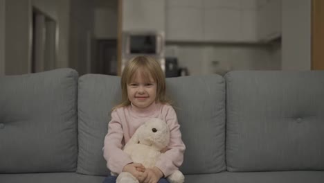 Pretty-happy-little-girl-in-casual-wearing-sitting-on-sofa-with-toy-dog-and-smiling