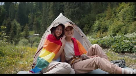 Porträt-Zweier-Lesbischer-Mädchen,-Die-Sich-In-Die-LGBT-Flagge-Gehüllt-Haben-Und-Neben-Einem-Zelt-Vor-Dem-Hintergrund-Eines-Grünen-Waldes-Sitzen.-Kampagne-Von-Menschen-Mit-Nicht-traditioneller-Sexueller-Orientierung