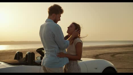 A-happy-blonde-girl-in-a-white-top-and-her-bearded-boyfriend-in-a-blue-shirt-are-stroking-each-other's-faces-and-hugging-near-a-white-convertible-car.-A-romantic-summer-meeting-against-the-river-and-the-yellow-sky