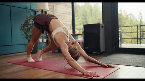 A-blonde-girl-with-tattoos-in-a-white-top-does-an-exercise-to-stretch-and-tone-the-muscles-of-the-back-and-shoulders-on-a-special-red-rug-in-a-house-overlooking-the-forest