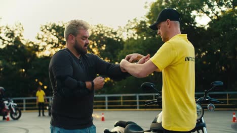 Driving-instructor-in-yellow-t-shirt-helping-a-biker-with-a-beard-to-put-on-elbow-protection.-Protective-clothing-as-an-important-part-of-a-motorcycle-trip
