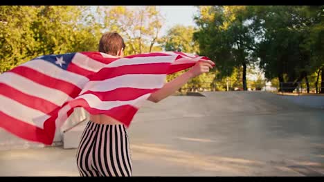 A-happy-short-haired-girl-in-a-purple-top-in-red-headphones-rides-in-a-skate-park-on-roller-skates-and-holds-the-USA-flag-in-her-hands