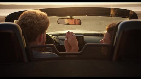 A-guy-and-his-girlfriend-are-sitting-in-a-convertible-car-and-holding-each-other's-hand-in-windy-weather-against-the-river-in-the-evening
