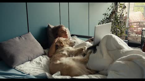 A-blonde-girl-lies-on-the-bed-with-her-dog-and-watches-a-movie-on-a-laptop-in-the-morning.-Holidays-with-pets