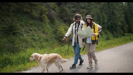 A-guy-and-a-girl-tourists-in-special-hiking-clothes,-together-with-their-light-colored-dog,-are-looking-at-a-map-in-order-to-understand-where-they-are-going-along-the-road-along-the-forest