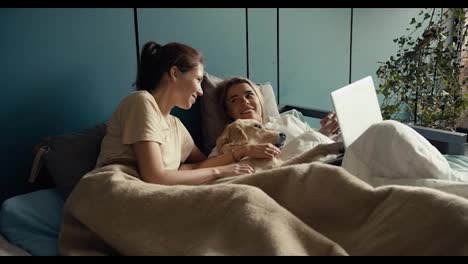 A-blonde-girl-and-a-brunette-girl-are-lying-in-bed-wrapped-in-a-blanket-with-their-light-colored-dog,-talking-and-looking-at-the-laptop-screen