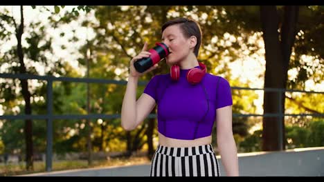 Portrait-of-a-girl-with-a-short-haircut-in-a-purple-top,-striped-pants-and-red-headphones-drinks-water-from-a-special-sports-bottle-in-a-skatepark-in-summer