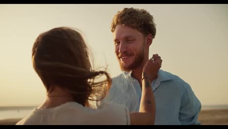 A-bearded-man-with-curly-hair-in-a-blue-shirt-is-talking-to-his-blonde-girlfriend,-who-is-stroking-his-chin-and-beard-against-the-yellow-sky-near-the-river