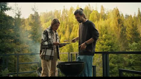 A-brunette-guy-in-a-brown-sweater-and-a-blonde-girl-fry-barbecue-meat-on-a-special-grill-on-the-balcony-of-a-country-house-in-a-coniferous-forest