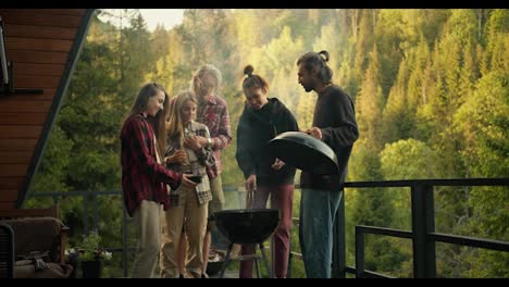 Friends-at-a-picnic:-a-girl-puts-sausages-on-a-wood-fired-grill,-her-friends-help-her-in-this.-Barbecue-on-the-balcony-of-a-country-house-in-the-mountains-with-a-coniferous-forest