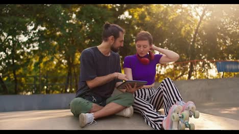 Brunette-guy-in-a-gray-t-shirt-explains-to-a-girl-with-a-short-haircut-in-a-purple-top-and-striped-pants-showing-an-electronic-tablet-in-a-skatepark-in-summer