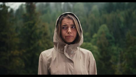 A-sad-blonde-girl-stands-against-the-background-of-forest-rain-in-the-mountains,-she-is-sad-and-looks-at-the-camera