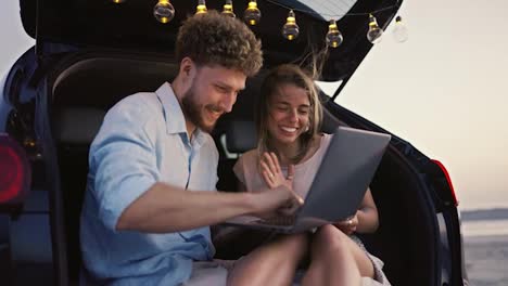 A-blonde-woman-waves-her-hand-at-a-laptop-screen-while-her-boyfriend-looks-at-a-laptop-screen-with-her-and-smiles-while-sitting