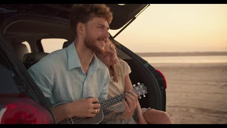 A-bearded-man-with-curly-hair-in-a-blue-shirt-is-playing-the-ukulele-and-singing,-while-his-blonde-girlfriend-is-resting-her-head-on-his-shoulder.-They-are-smiling-and-sitting-in-the-trunk-of-the-black-car-against-the-yellow-sky