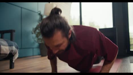 A-happy-brunette-guy-in-a-red-t-shirt-has-done-the-push-up-exercise-at-home,-gets-up-on-his-feet-and-wipes-his-forehead.-Fitness-at-home