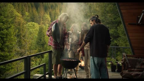 A-group-of-friends-at-a-picnic:-a-guy-in-a-red-shirt-lifts-the-lid-of-the-barbecue-and-thick-white-smoke-comes-out.-Grilling-food-at-a-picnic-with-mountain-and-forest-view