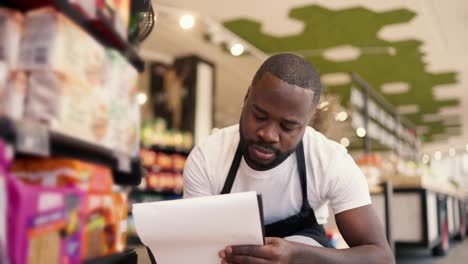 Portrait-of-a-man-with-Black-skin-color-in-a-white-t-shirt-and-a-black-apron-sat-down-near-the-shop-window-and-writes-down-the-goods-on-paper-on-the-tablet