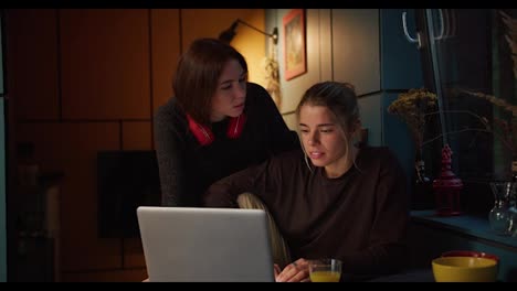 A-brunette-girl-and-a-blonde-girl-have-fun-and-look-at-the-screen-of-a-white-laptop-in-a-dark-room,-with-a-sacred-yellow-lamp,-in-the-evening