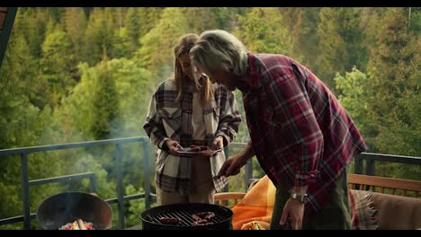 A-blonde-girl-helps-her-blonde-boyfriend-remove-sausages-from-the-grill-on-the-balcony-of-a-country-house-overlooking-the-forest-and-mountains