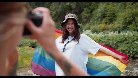 POV-shot-of-a-girl-taking-a-picture-of-her-friend-who-is-standing-with-an-LGBT-flag-in-a-white-T-shirt-against-the-backdrop-of-a-green-forest