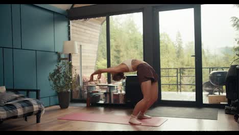 A-blonde-girl-in-a-white-top-does-stretching-exercises,-she-bends-her-back-and-stands-on-her-hands-in-an-industrial-house-overlooking-a-green-forest.-bridge-exercise