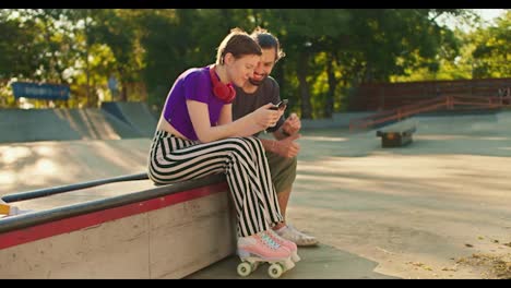 Side-view-of-a-girl-with-a-short-haircut-in-a-purple-top-striped-pants-in-pink-roller-skates-sits-on-a-bench-in-a-skate-park-and-communicates-with-a-brunette-guy-in-a-gray-t-shirt-with-a-phone-in-her-hands