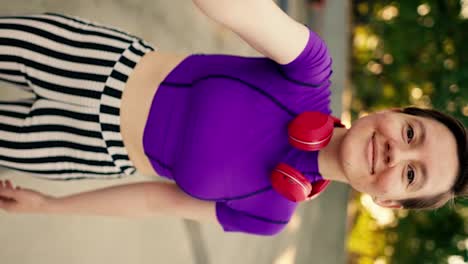Vertical-video:-Happy-girl-with-short-haircut-in-purple-top-red-headphones-and-striped-pants-rides-in-a-skate-park-on-roller-skates-and-takes-a-selfie-and-shows-V-sign