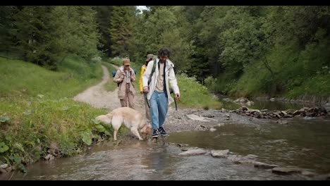 A-guy-and-two-girls-in-hiking-clothes,-together-with-their-dog,-are-trying-to-cross-a-mountain-river-along-a-special-path-made-of-stones