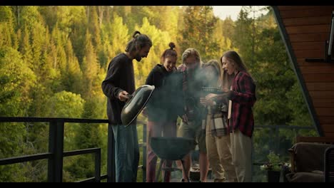 Five-friends-cook-food-on-the-grill-on-the-balcony-of-a-country-house-overlooking-the-mountains-and-green-forest.-Friends-chatting-and-laughing-at-a-picnic