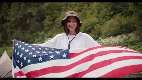 Portrait-of-a-brunette-girl-in-a-white-T-shirt-who-stands-with-the-flag-of-the-United-States-of-America-that-flutters-in-the-wind-against-the-background-of-a-green-forest
