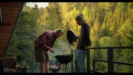 A-blond-guy-in-a-red-plaid-shirt-turns-sausages-on-the-grill,-and-his-friend,-a-brunette-guy,-tells-you-how-to-do-it-right.-Friends-on-a-picnic-overlooking-the-coniferous-forest-and-mountains