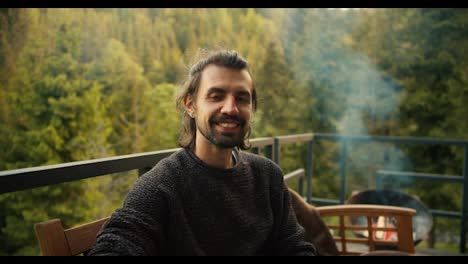 A-young-brunette-man-in-a-green-sweater-poses-at-a-picnic-overlooking-the-mountains-and-coniferous-forest.-Portrait-of-a-happy-man-resting-outside-the-city