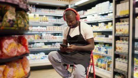 a-man-with-Black-skin-color-in-red-headphones-and-a-black-apron-sits-on-a-chair-and-listens-to-music-in-a-supermarket.-Break-at-work