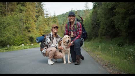 A-guy-and-a-girl-in-hiking-clothes,-together-with-their-light-colored-dog,-pose-and-look-at-the-camera-while-standing-near-the-road-against-the-backdrop-of-the-forest