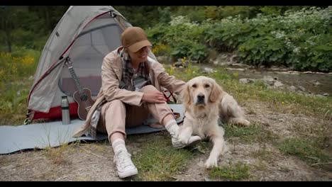 Portrait-of-a-blonde-girl-in-camping-clothes,-who-sits-near-her-tent-and-strokes-her-light-colored-dog-against-the-background-of-a-green-forest-and-a-mountain-river